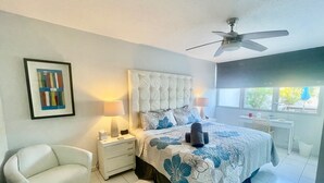 A bedroom with a tufted headboard, two bedside lamps, a bed with floral bedding, a white armchair, and a ceiling fan. - MB2E-Unique Terrace Two Bedrooms-Pool & Ocean View (Carolina)