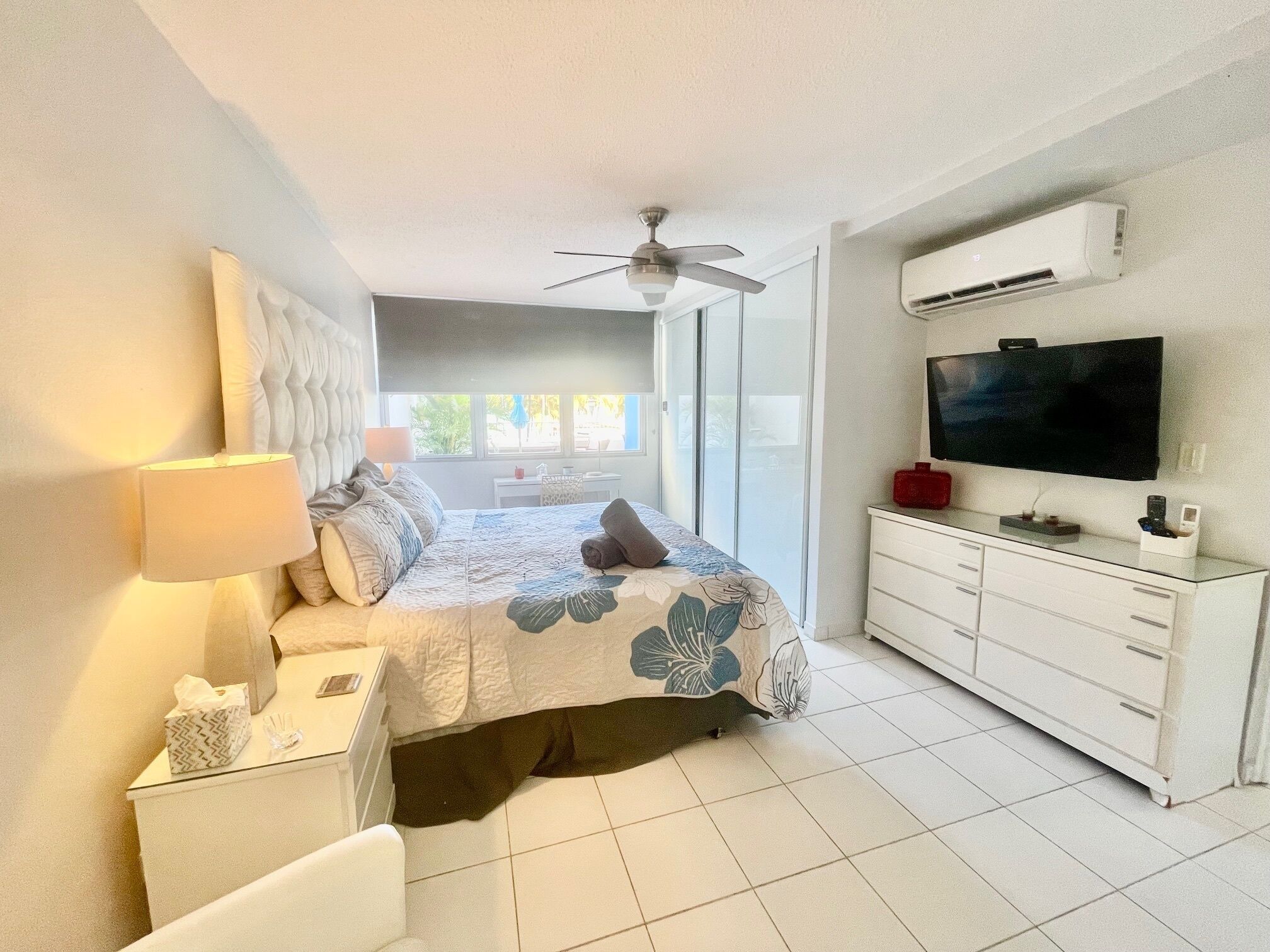 Quarto com cama, mesa de cabeceira, abajur, televisão e ventoinha no teto.