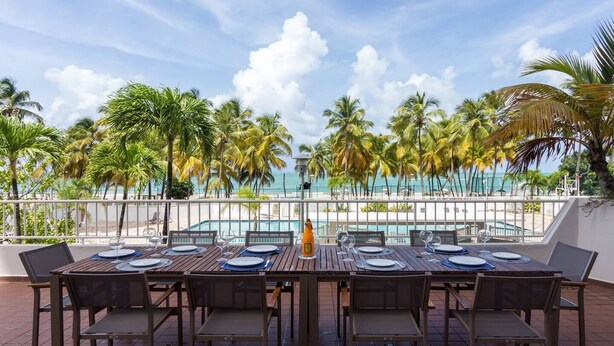 An outdoor dining area with tables and chairs, overlooking a pool and palm trees, with a view of the ocean. - MB2E-Unique Terrace Two Bedrooms-Pool & Ocean View (Carolina)