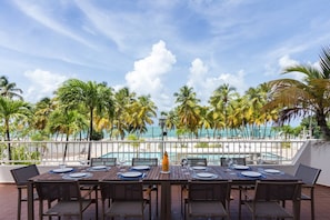 An outdoor dining area with tables and chairs, overlooking a pool and palm trees, with a view of the ocean.