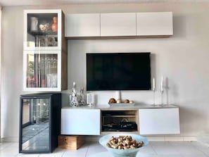 A modern living room with a flat-screen TV mounted above a white console, a glass-fronted cabinet with decorative items, and a small glass table with a bowl of snacks.