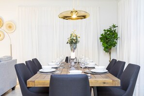 A dining area with a wooden table set for six, surrounded by dark chairs, with a pendant light above and a potted plant to the side.