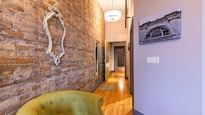 A hallway with a brick wall, a green upholstered chair, a wooden floor, a door, and a wall-mounted light fixture. - 124 Lofts #3 - Downtown Dundee - Walk to Dining (West Dundee)
