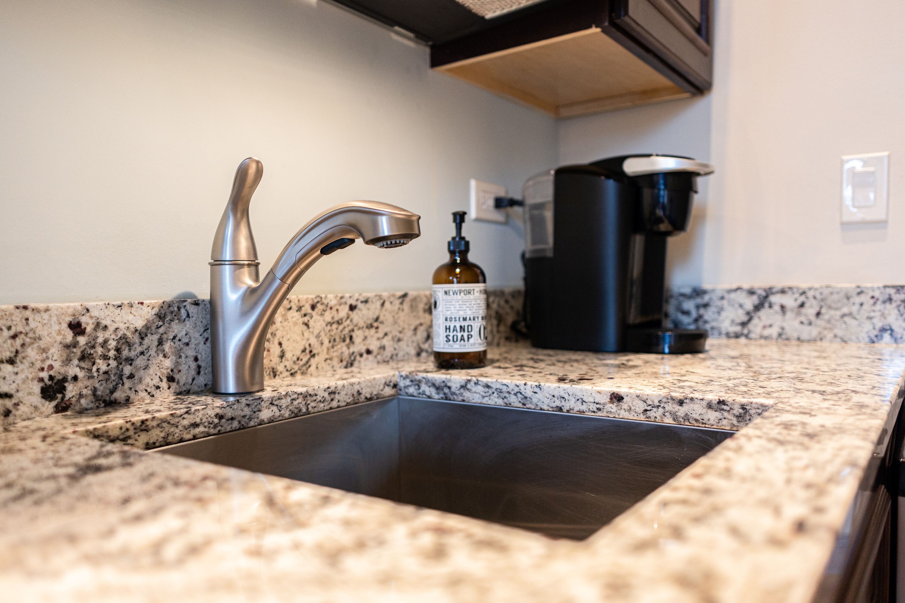 A kitchen with a granite countertop, a stainless steel sink, a coffee maker, and a bottle of hand soap.