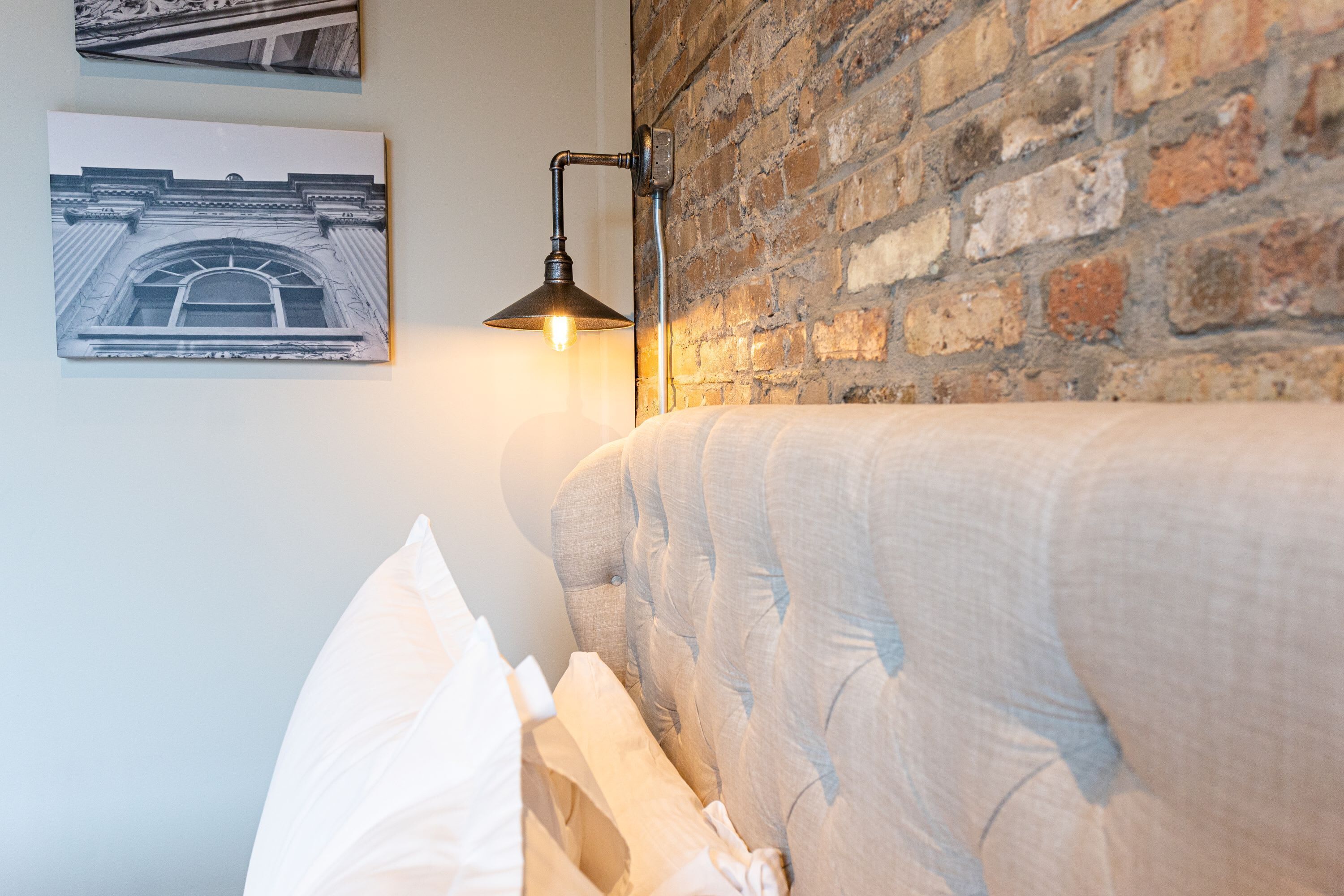 A bed with a headboard against a brick wall, a wall-mounted lamp, and a framed picture above the bed.