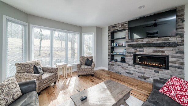 A living room with a fireplace, a TV mounted on a stone wall, and a large window with blinds. - Beautiful 4-season Mountain Home- New Listing (Wintergreen Resort)