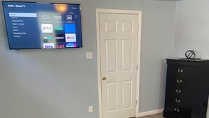 A living room with a flat-screen TV mounted on the wall displaying a streaming service interface, a door, a black cabinet, and a patterned rug.