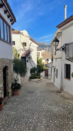 A cobblestone street in a historic town with white buildings, stone arches, and potted plants.