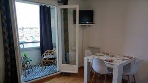 Une pièce comprenant une table à manger dressée pour deux personnes, un canapé, un téléviseur fixé au mur et une vue sur une marina, visible à travers une porte-fenêtre coulissante.