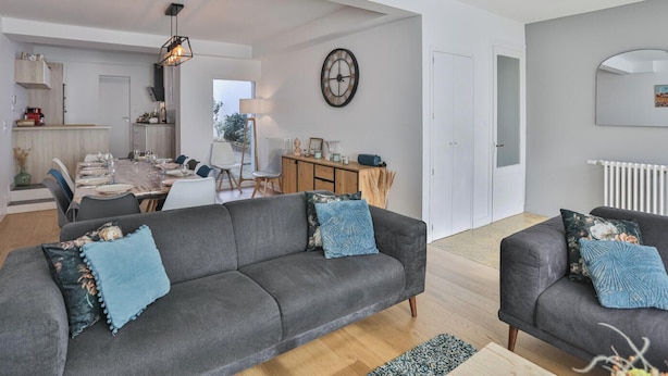 Ein modernes Wohnzimmer mit grauem Sofa, einem Essbereich und einer Küche im Hintergrund.