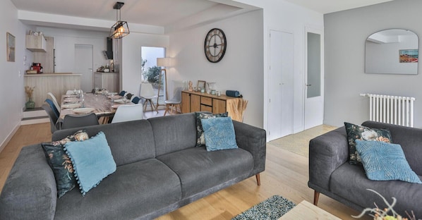 A modern living room with a grey sofa, a dining area with a table and chairs, a clock on the wall, and a radiator.