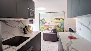 Una cocina compacta con encimera de mármol, estufa integrada y un sofá gris en la zona de estar contigua. Se observa un cuadro grande de un paisaje en la pared.