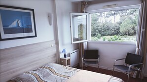 Un dormitorio con cama, una silla, un escritorio, una lámpara y ventana con vistas a la vegetación.