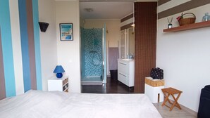 Ein Schlafzimmer mit einem Bett, einem Nachttisch mit Lampe, einem Koffer, einem kleinen Holzschemel und einem Badezimmer mit Dusche und Waschbecken.