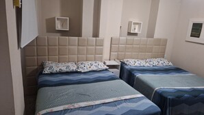 A hotel room with two beds, each with a headboard and floral-patterned bedding. There are wall-mounted light fixtures and a small shelf with a decorative item. - Pelourinho, complete house. (Salvador)