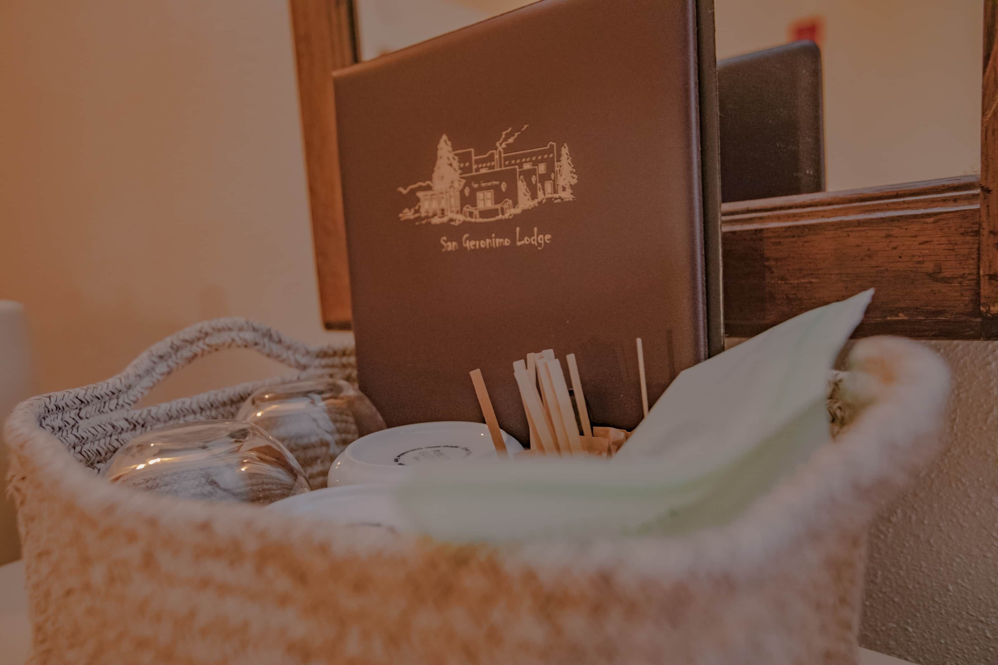 A brown booklet with 'San Geronimo Lodge' printed on it, placed on a wooden surface next to a woven bag containing a bottle, a glass, and a white object.