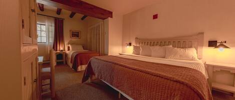 A bedroom with a large bed, a smaller bed, and a desk. There is a window with curtains, a wooden ceiling beam, and a wall-mounted light.