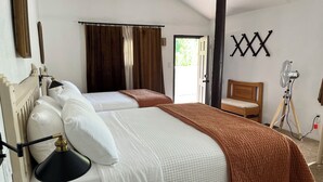 Camera d'albergo con due letti, una testiera in legno, una lampada da comodino, una ventola e una porta che dà su un balcone.