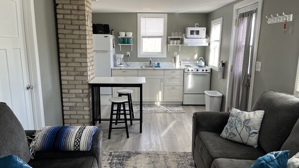 A compact kitchen with a brick accent wall, a white refrigerator, a microwave, and a stove. There are two bar stools, a gray sofa with blue pillows, and a patterned rug. - Small Home in the City of Alexandria! (Alexandria)