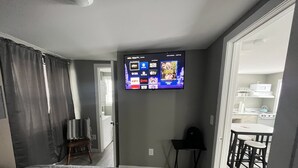 A hotel room with a bed, a TV mounted on the wall displaying various channels, a small table with two chairs, and a view of the kitchen through an open door. - Small Home in the City of Alexandria! (Alexandria)