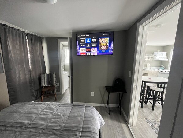 Une chambre d’hôtel avec un lit, un téléviseur fixé au mur diffusant différentes chaînes, une petite table avec deux chaises, et une vue sur la cuisine par une porte ouverte.