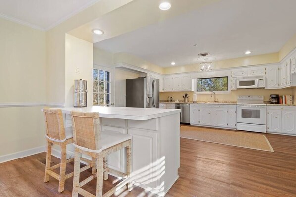 A kitchen with white cabinets, a central island, and a dining area with wicker chairs. - HIGH TIDE: Spacious Comfort Next to Tides Inn (Irvington)