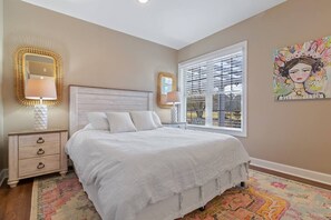 A bedroom with a bed, two bedside lamps, a nightstand, a window with blinds, and a colorful rug.