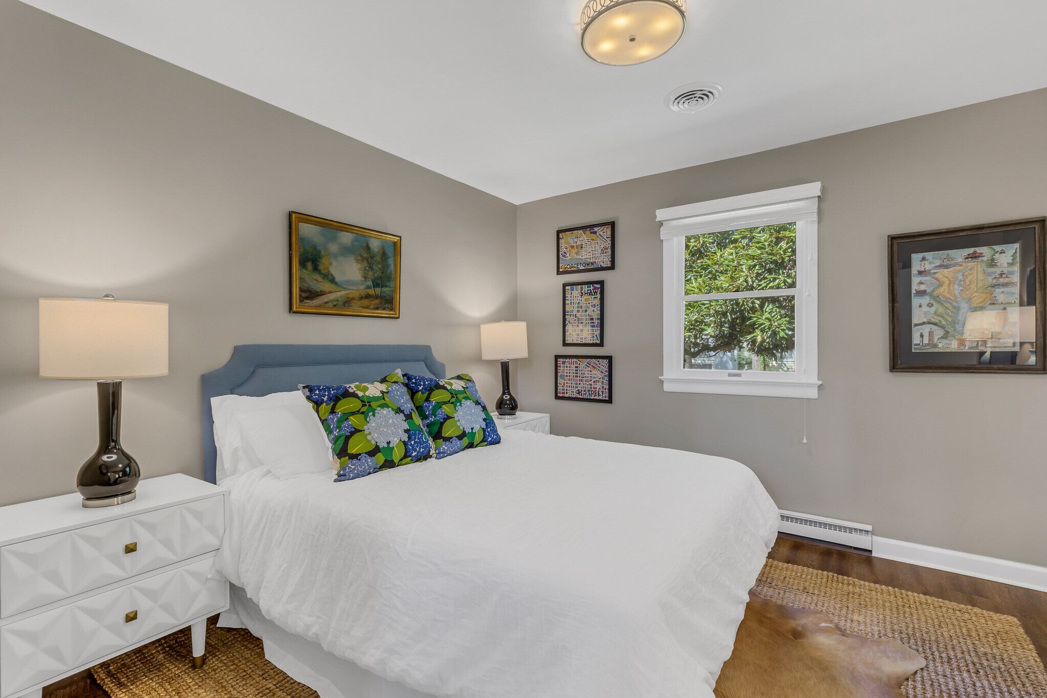 A bedroom with a bed, two bedside tables with lamps, a window with a view of greenery, and framed artwork on the walls.