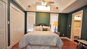 A bedroom with a bed, a nightstand, a lamp, a bench, and a bathroom visible through an open door.