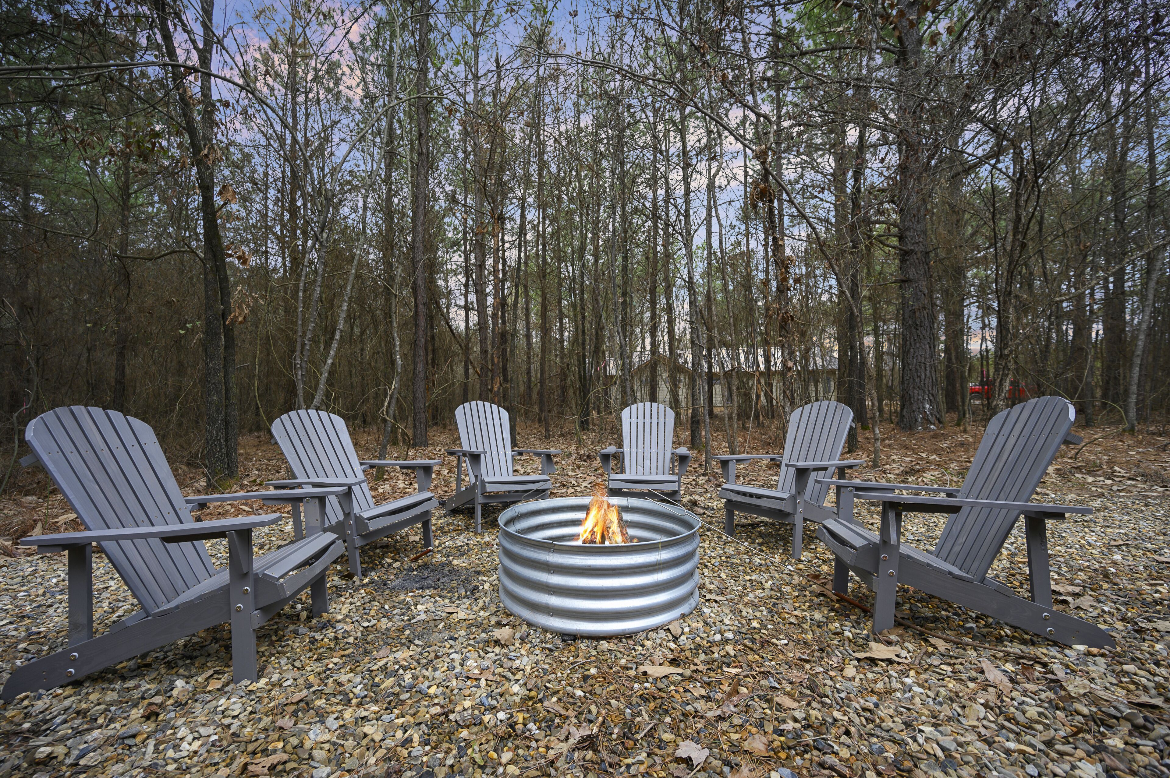 A circular metal fire pit with four Adirondack chairs arranged around it in a forested area.
