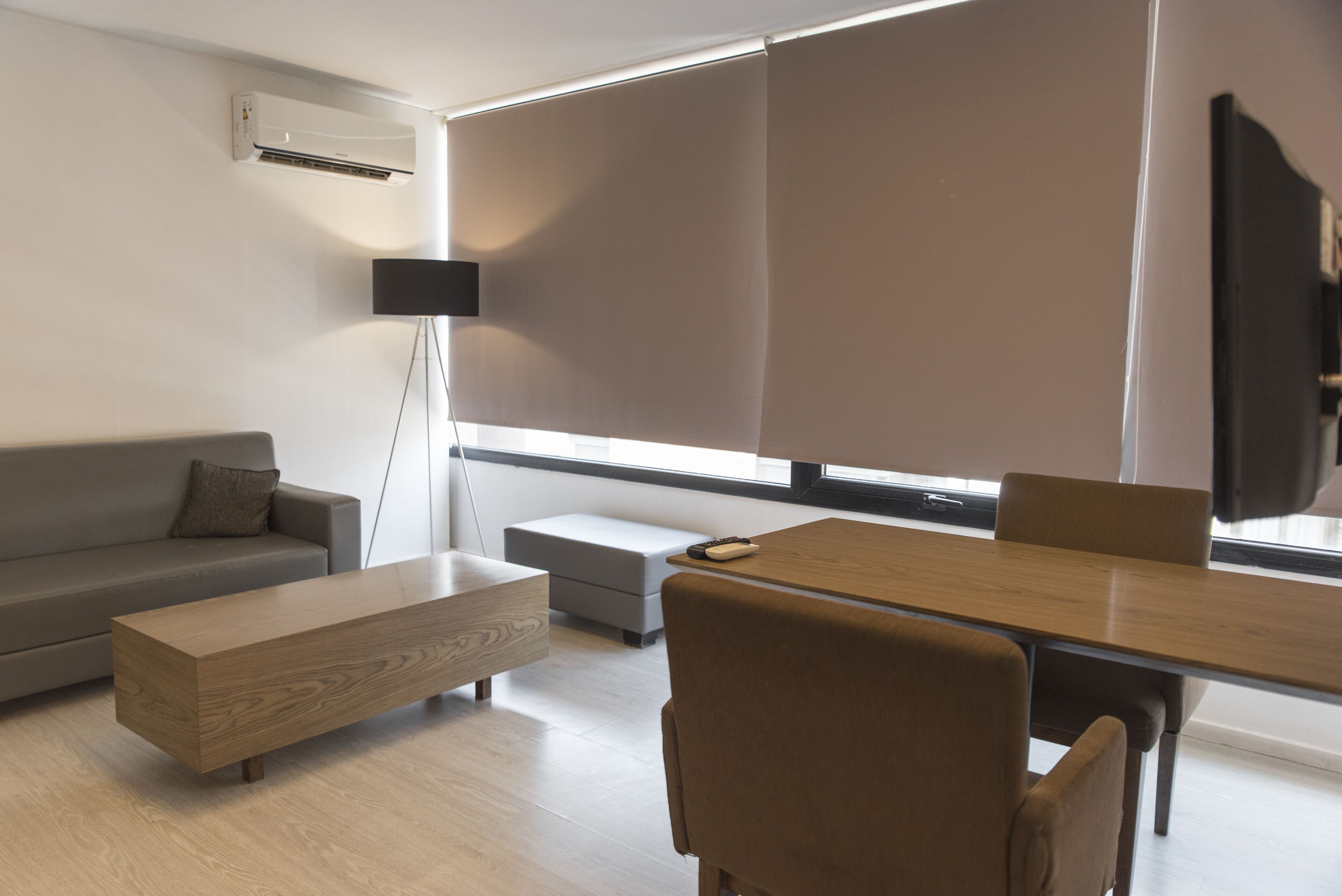 A modern living room with a sofa, a wooden coffee table, a dining table with chairs, a floor lamp, and a large window with blinds.