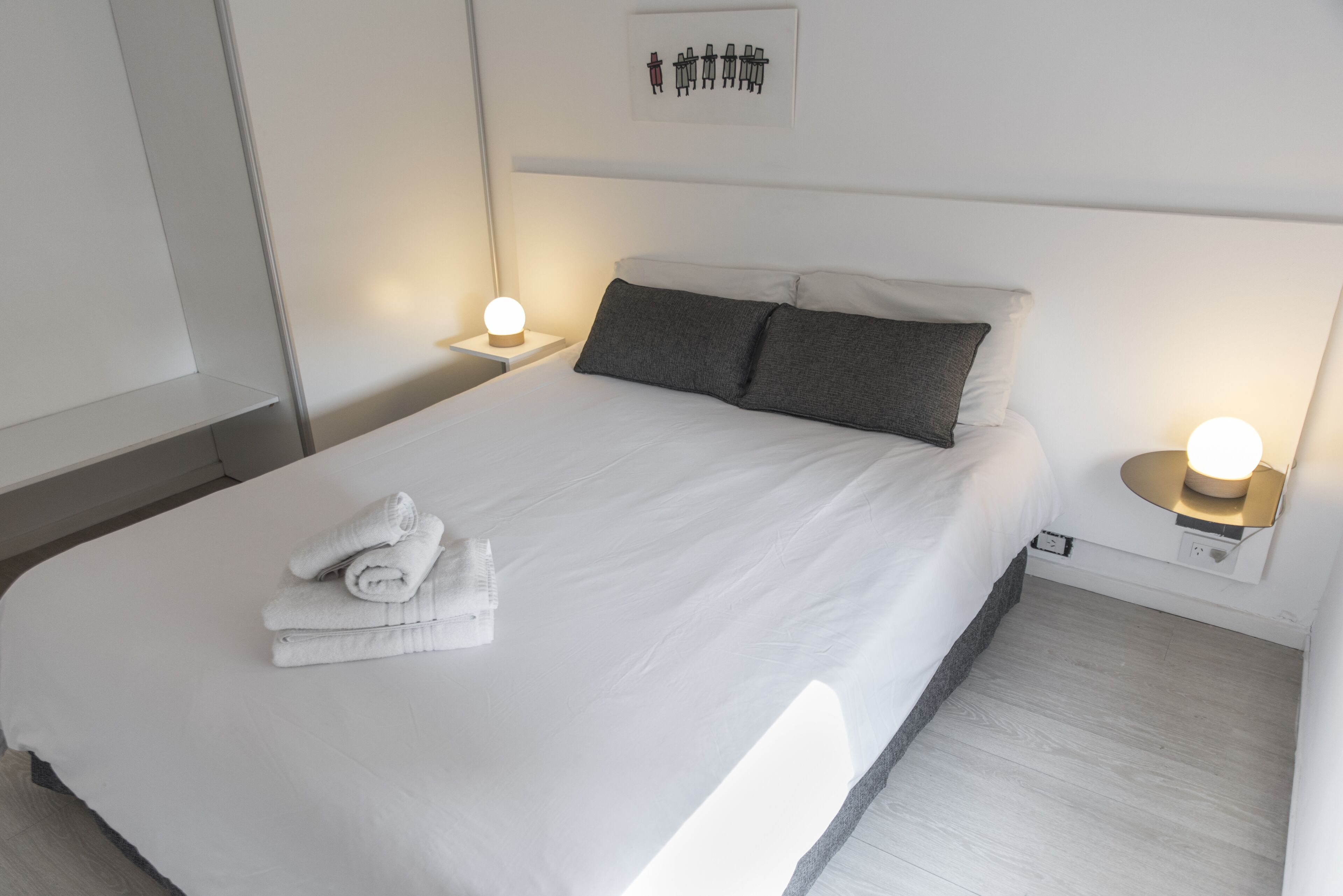 A neatly made bed with white linens and two pillows, a small bedside table with a lamp, and a wall-mounted artwork above the bed.