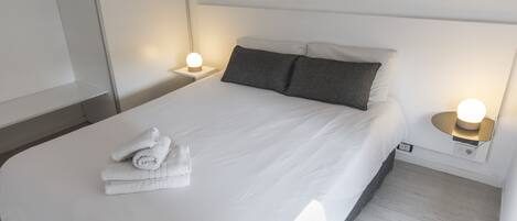 A neatly made bed with white linens and two pillows, a small bedside table with a lamp, and a wall-mounted artwork above the bed.