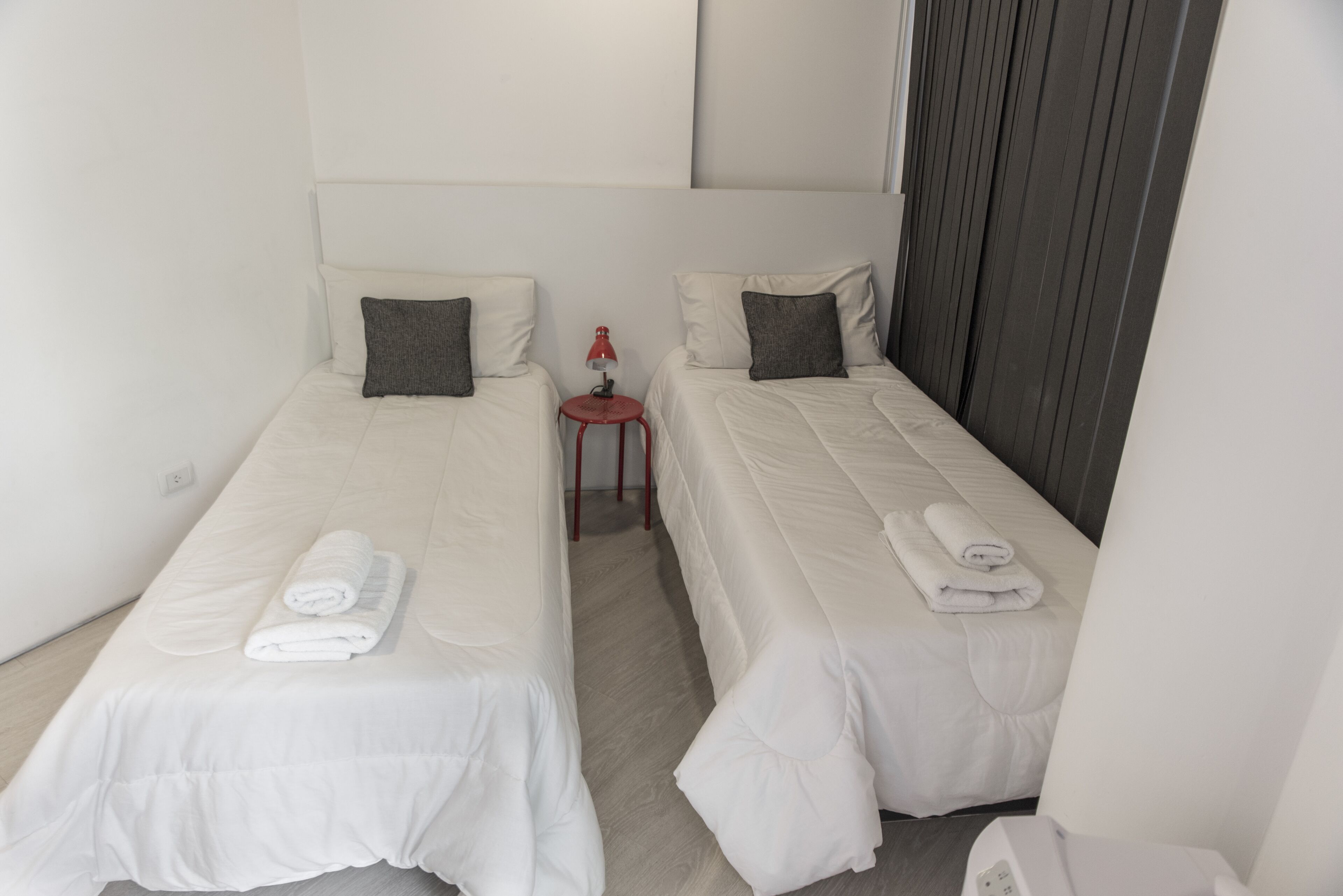 Habitación de hotel con dos camas individuales, cada una con una almohada y una toalla enrollada. Hay una pequeña mesa con una lámpara roja entre las camas.