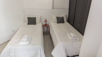 Habitación de hotel con dos camas individuales, cada una con una almohada y una toalla enrollada. Hay una pequeña mesa con una lámpara roja entre las camas.