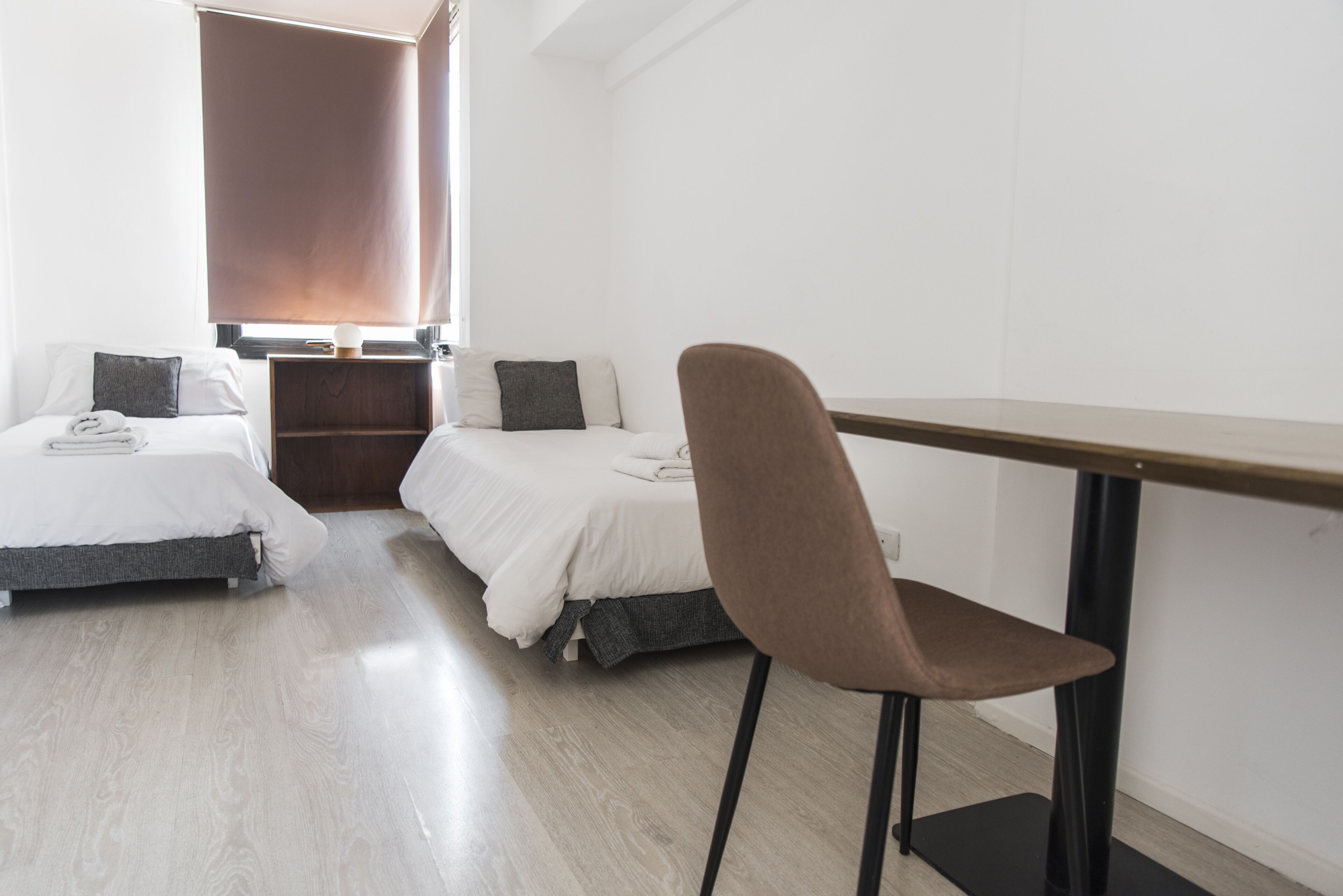 Una habitación con dos camas, una silla, una mesa y un ventanal con persianas.
