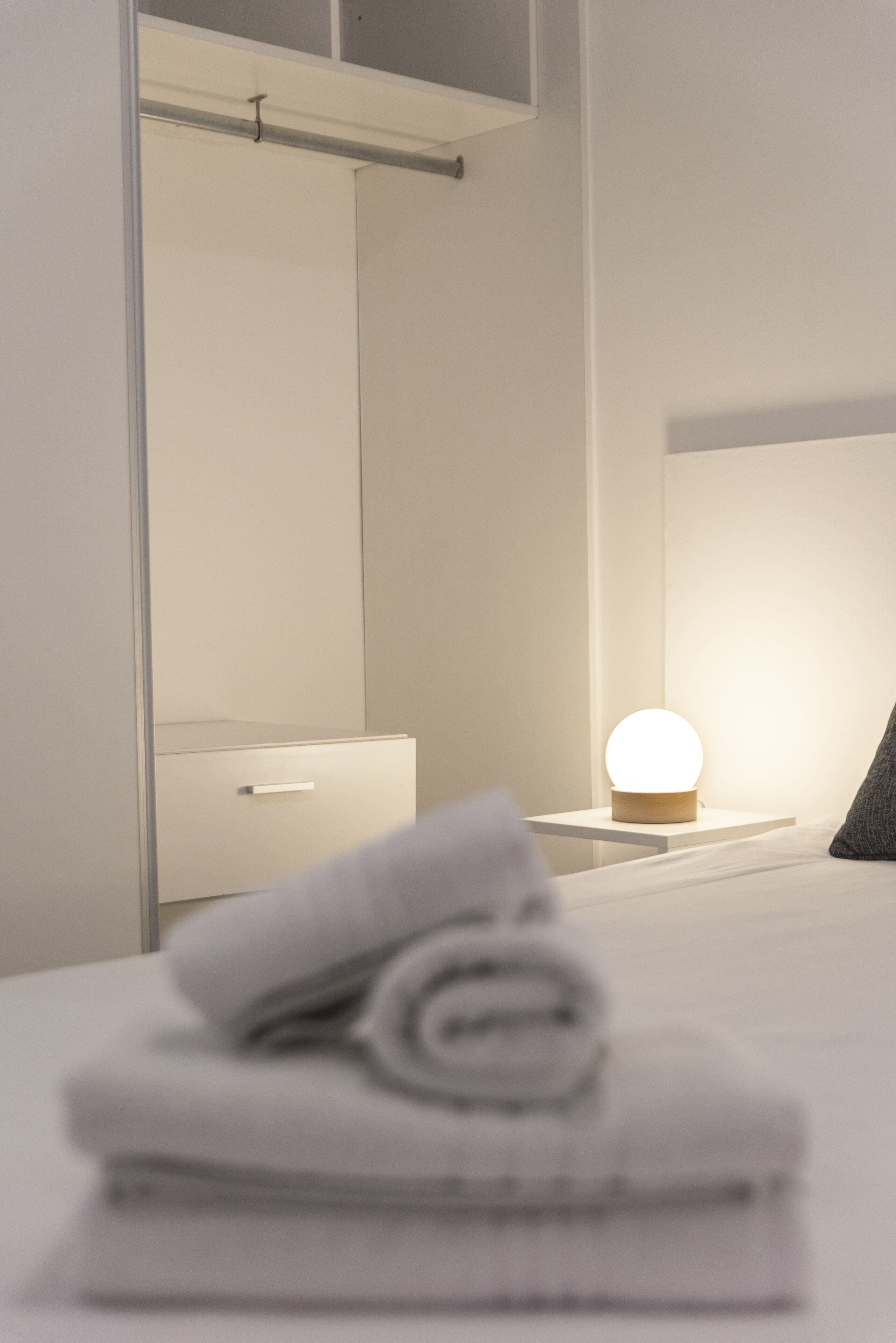 A neatly made bed with a folded white towel, a bedside table with a round light, and a mirror above a white dresser.
