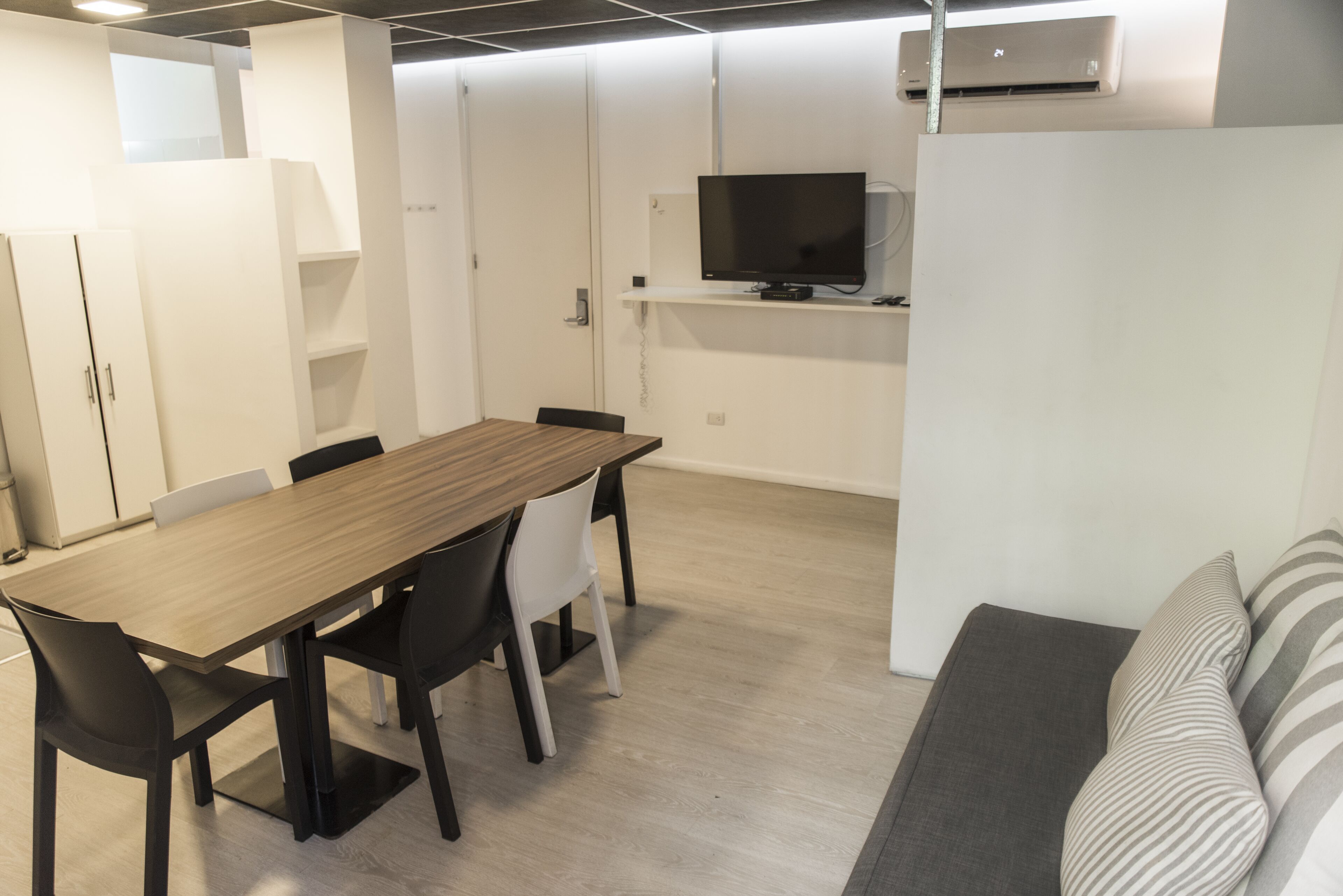 A modern, minimalist room with a wooden dining table, black chairs, a grey sofa, and a flat-screen TV mounted on a white wall.