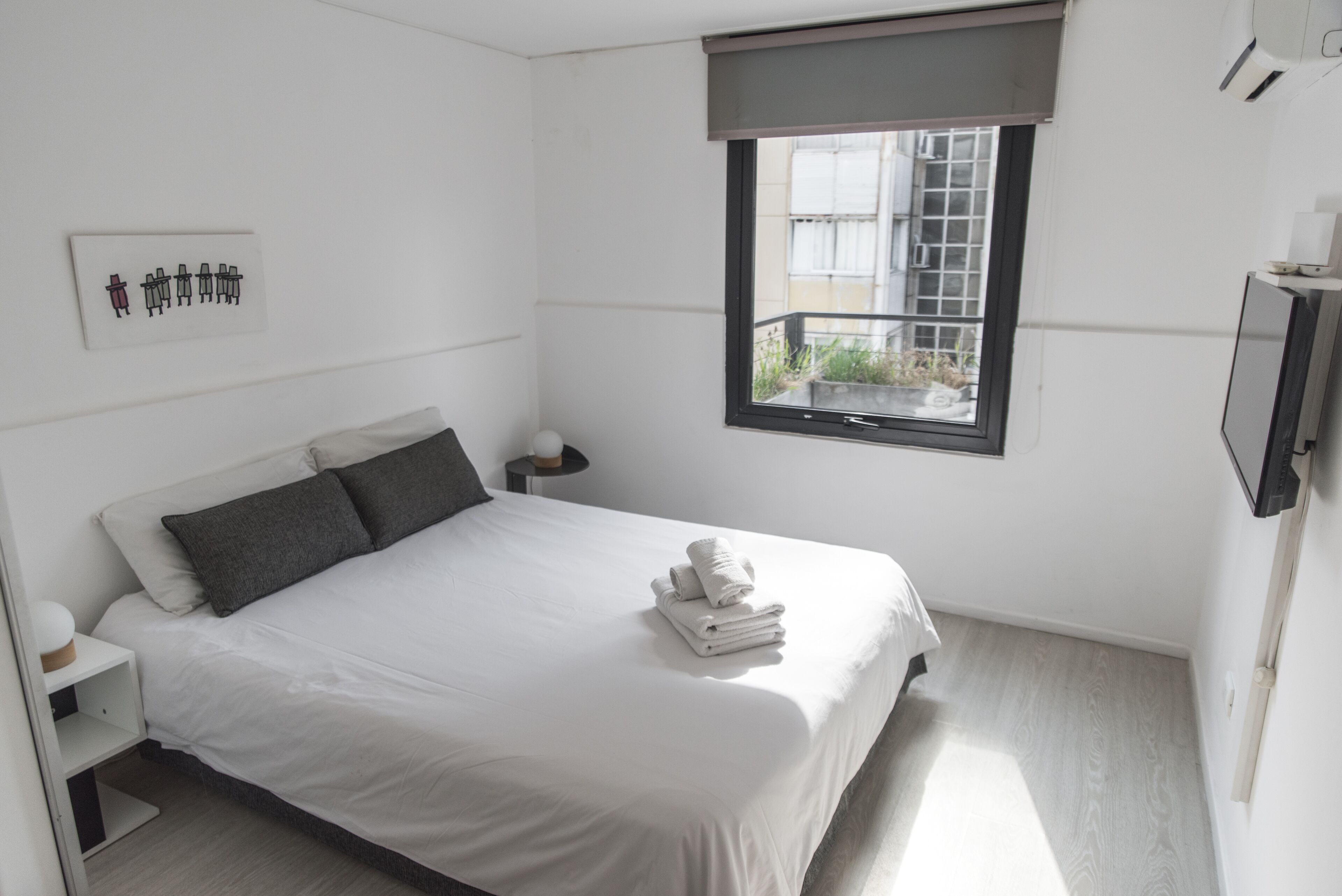 Una cama bien hecha con edredón blanco y almohadas, una mesita de noche con lámpara, una ventana con vista a edificios y un televisor montado en la pared.