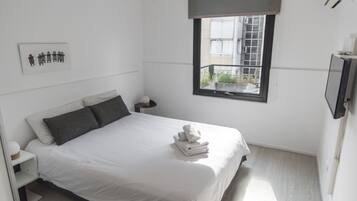 Una cama bien hecha con edredón blanco y almohadas, una mesita de noche con lámpara, una ventana con vista a edificios y un televisor montado en la pared.