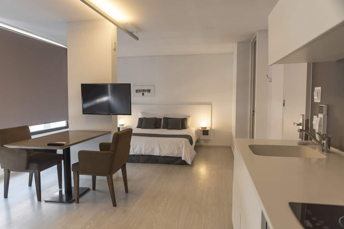 Habitación de hotel moderna con cama, mesita de noche, lámpara, televisor y una kitchenette con fregadero y estufa.