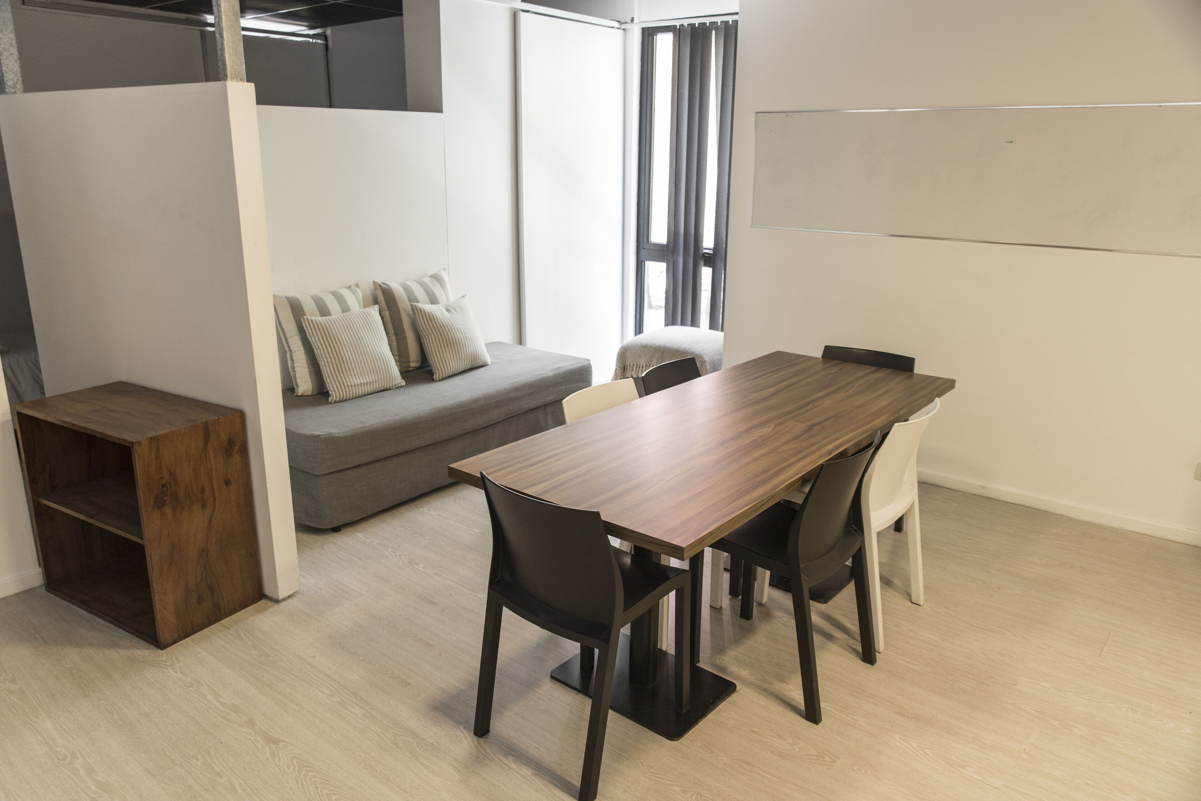 A modern, well-lit room with a wooden dining table, black chairs, a grey sofa with striped cushions, and a whiteboard on the wall.