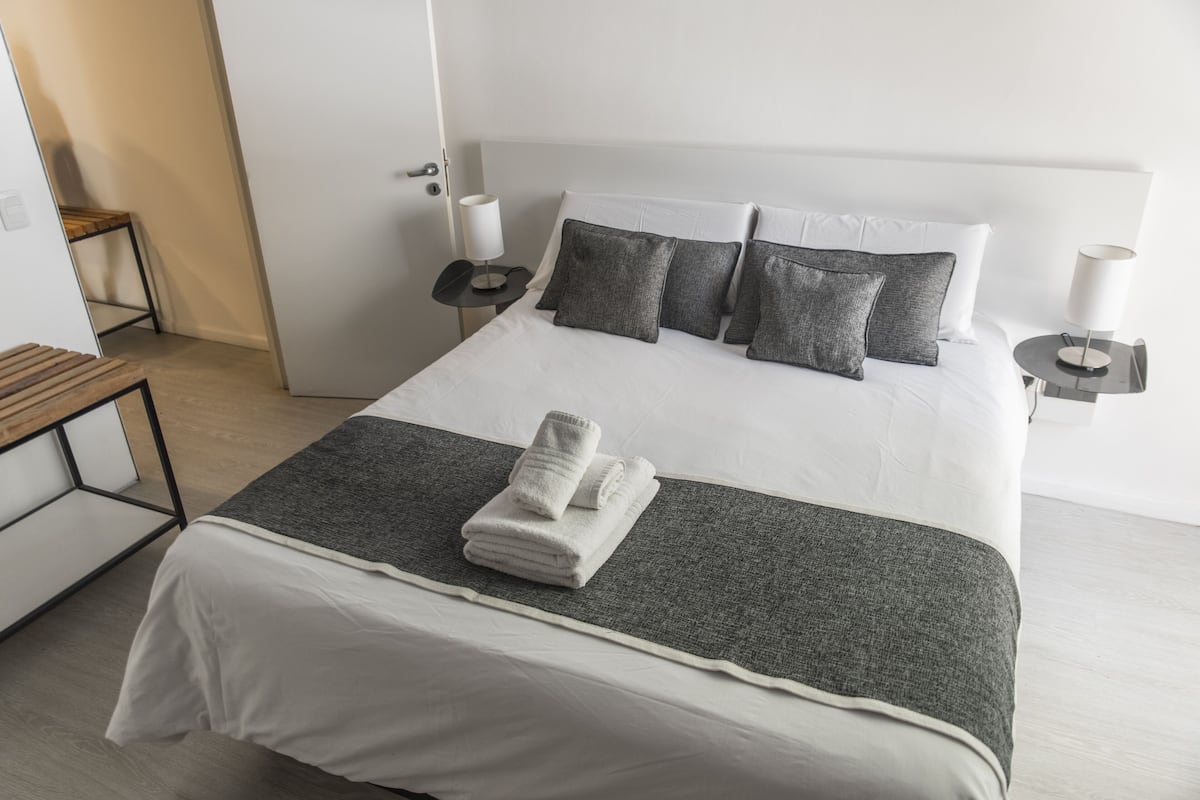 Una cama bien hecha con ropa de cama blanca y gris, una manta gris y toallas dobladas sobre la cama.