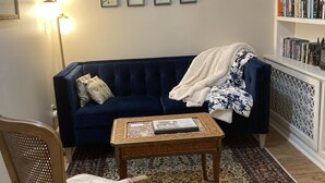 A cozy living room with a dark blue sofa, a patterned armchair, a small wooden coffee table, and a bookshelf filled with books. - Elegant Garden-Level Apt, Beautiful Marble Spa Bath, City Center! 96 Walk Score! (Denver)