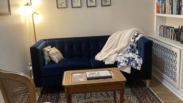 Een gezellige woonkamer met een donkerblauwe bank, een fauteuil met patroon, een klein houten bijzettafeltje en een boekenplank vol boeken.