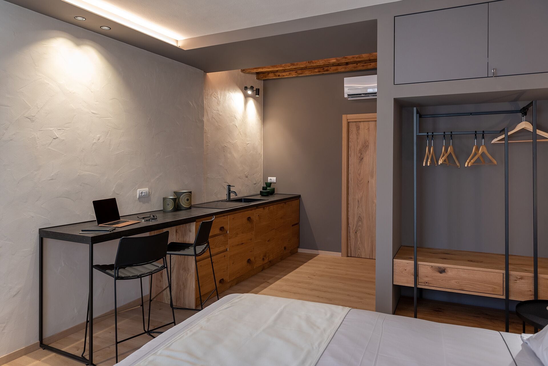 A modern bedroom with a bed, a desk with a chair, a small kitchenette, and a walk-in closet with wooden hangers.