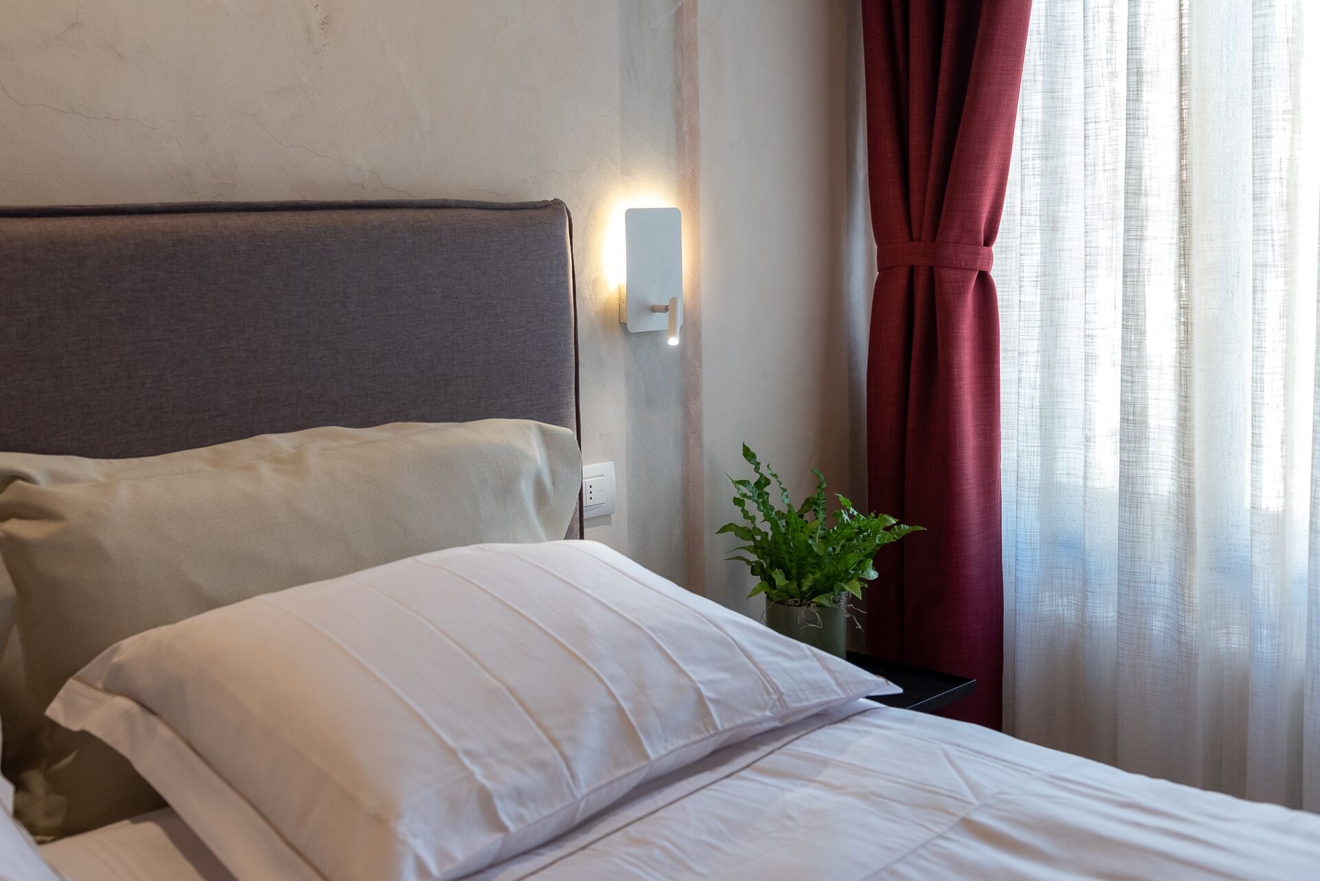 A hotel room with a bed, a nightstand, a wall-mounted light, a potted plant, and a window with curtains.