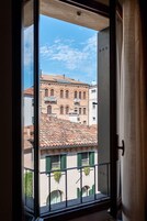 Vista a través de una ventana de un edificio con tejado de tejas, ventanas arqueadas y un balcón con plantas colgantes.