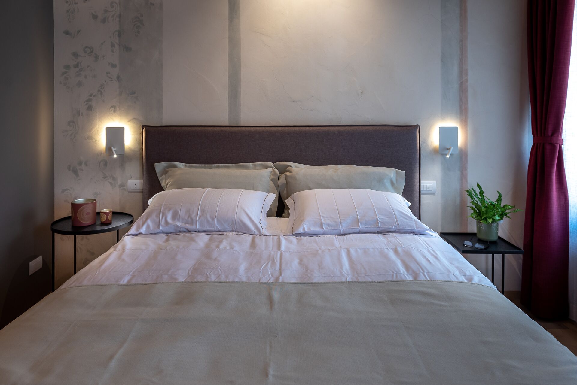 A neatly made bed with white linens, two bedside tables with lamps, a potted plant, and a window with curtains.