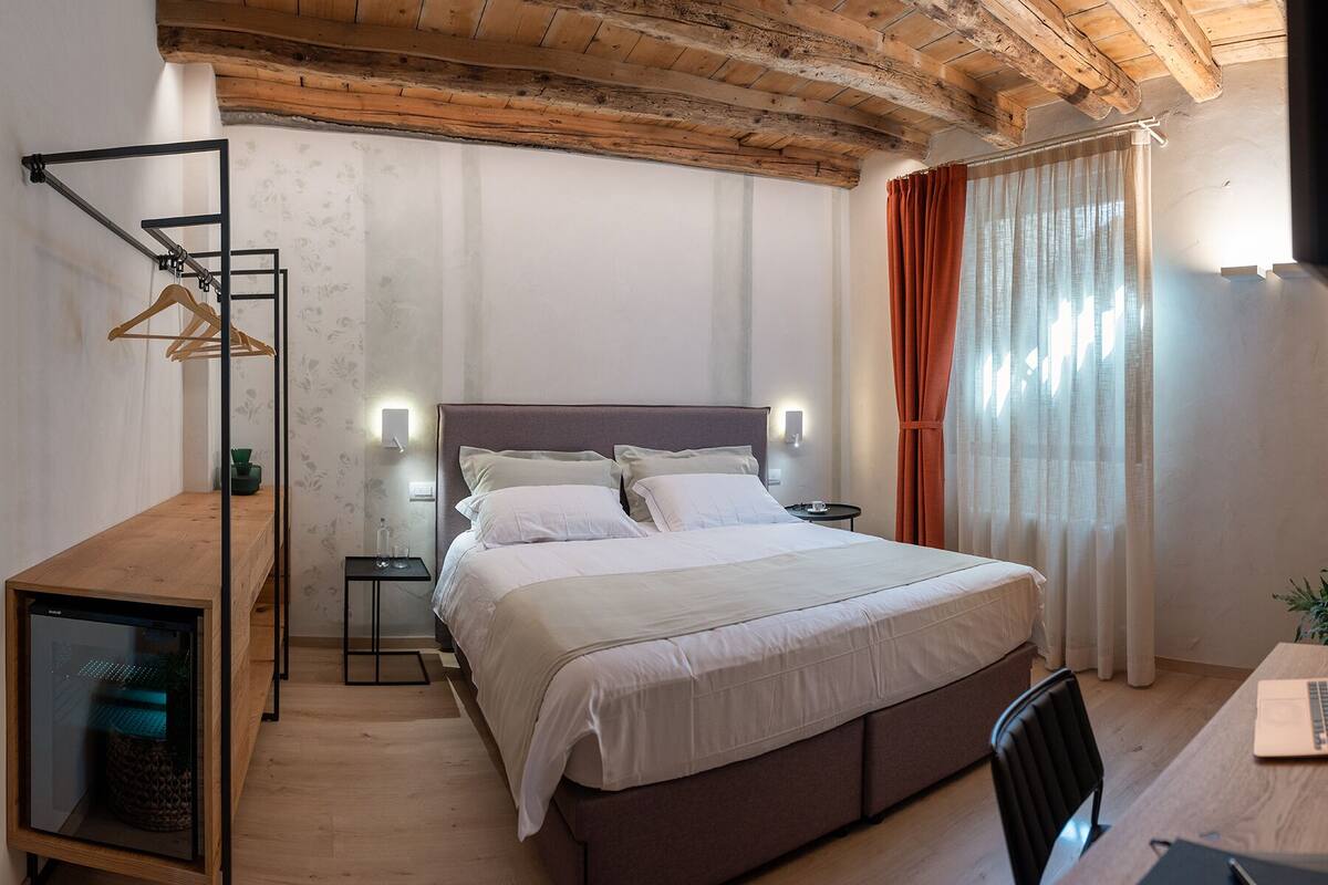 A bedroom with a bed, bedside tables, a desk with a laptop, a wooden floor, and a ceiling with exposed beams.
